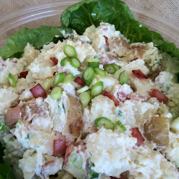 A bowl of creamy potato salad with spring onions and bacon bits on a bed of lettuce.