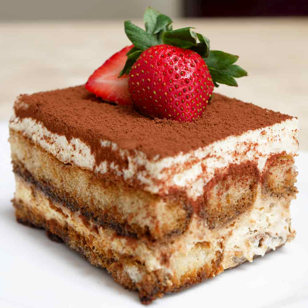 A square slice of sponge based cake dusted with chocolate with two strawberries on top.